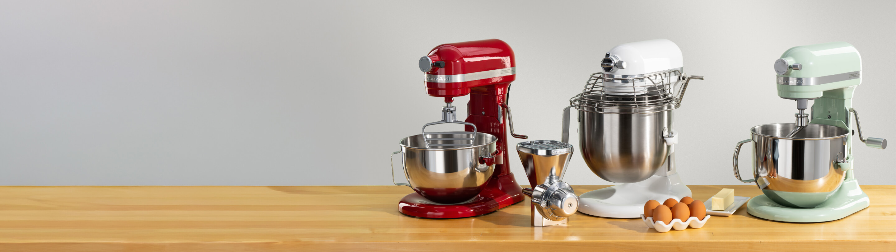 A trio of bowl-lift stand mixers in red, white and blue surrounded by different attachments and eggs on a wooden counter.