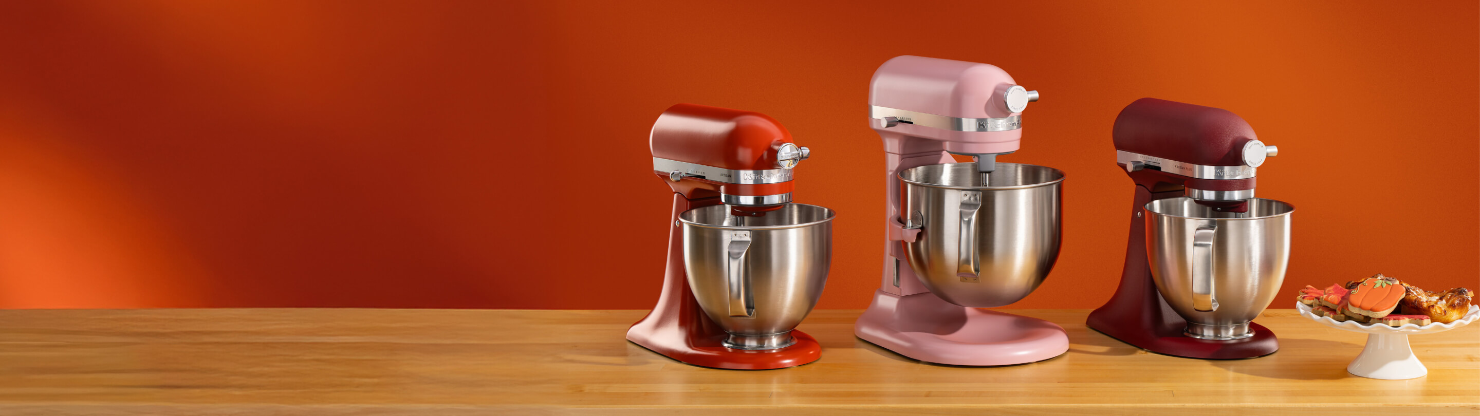 A red stand mixer, pink bowl-lift stand mixer and dark red stand mixer on a wooden counter with cookies on a cake platter