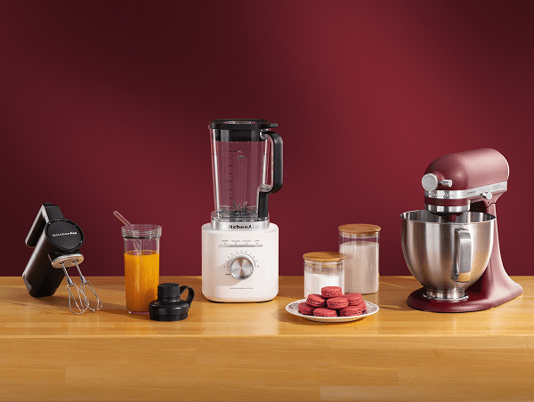 Black KitchenAid® hand mixer, white blender, and burgundy stand mixer on a wood counter beside macarons