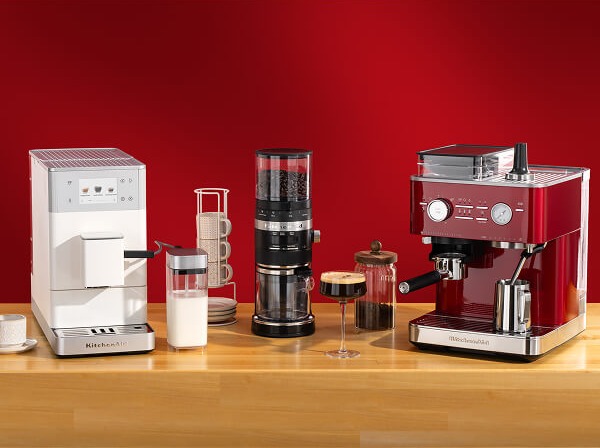 White KitchenAid® espresso machine, coffee grinder, and red espresso machine on a wooden counter with mugs and glassware
