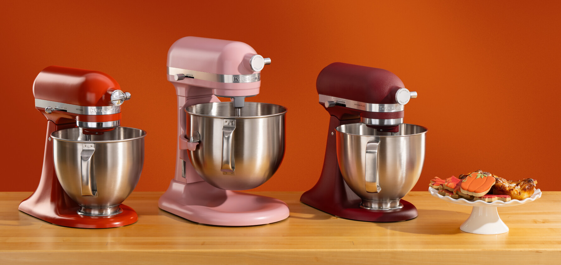 A red stand mixer, pink bowl-lift stand mixer and dark red stand mixer on a wooden counter with cookies on a cake platter