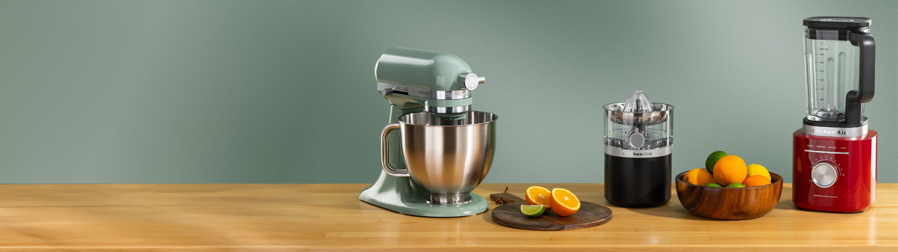 Green KitchenAid® stand mixer, black citrus juicer and red blender on a wooden counter with oranges
