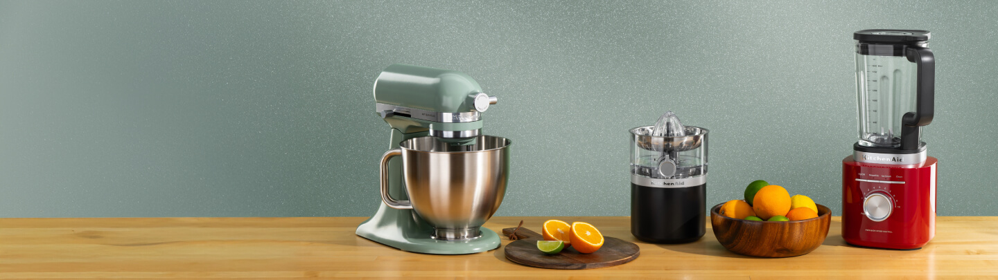 Green KitchenAid® stand mixer, black citrus juicer and red blender on a wooden counter with oranges