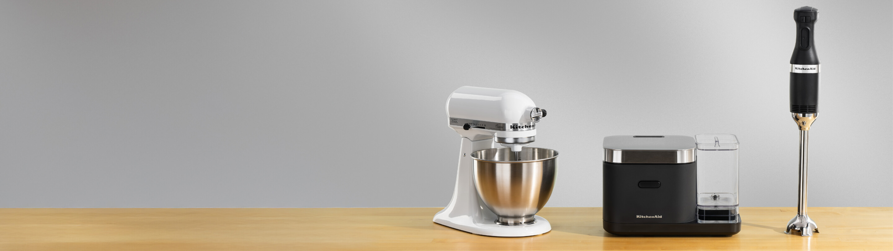 A white KitchenAid® stand mixer, a black grain and rice cooker and a black immersion blender on a wooden counter.