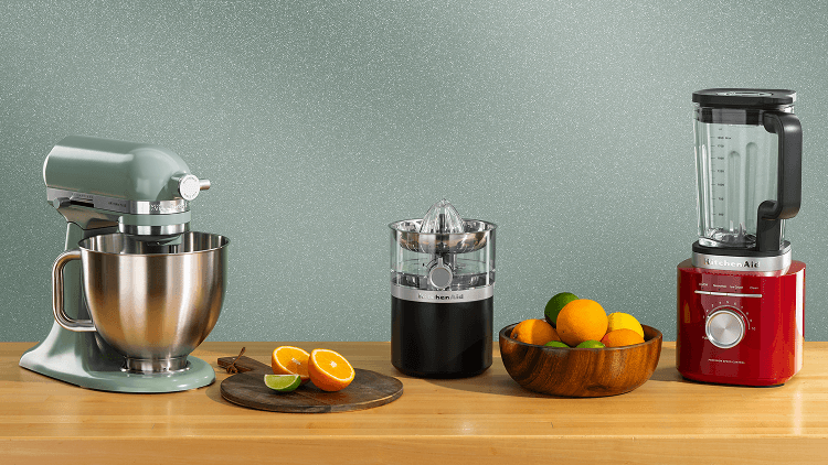 A teal stand mixer, black citrus juicer and red blender on a wooden counter with assorted citrus