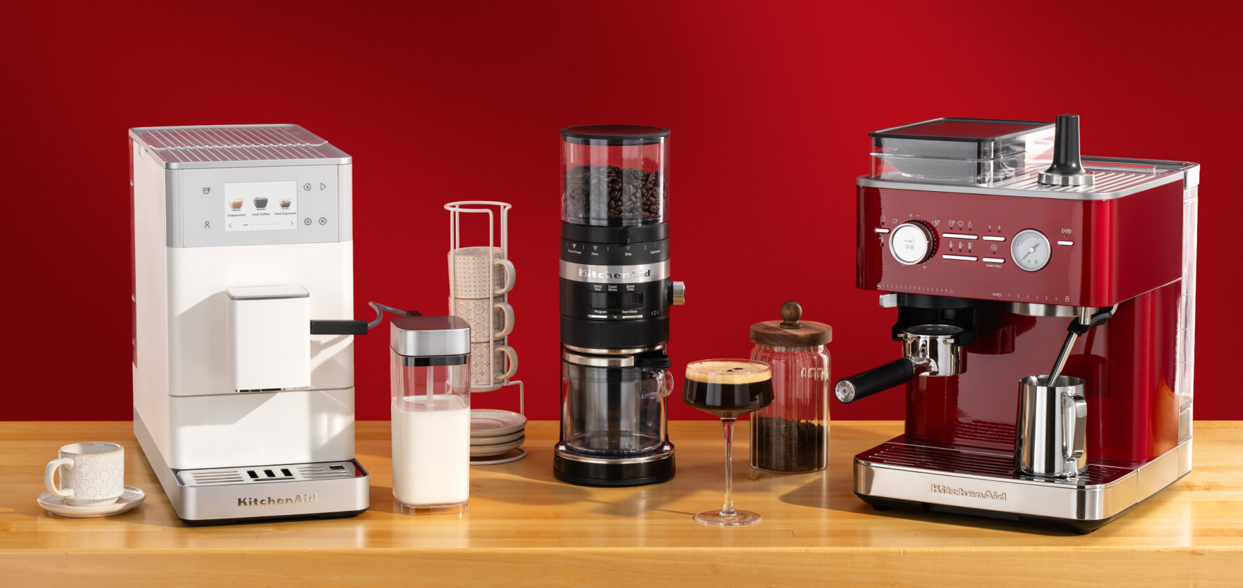 A white automatic espresso machine, black bean grinder and red espresso machine on a wooden countertop