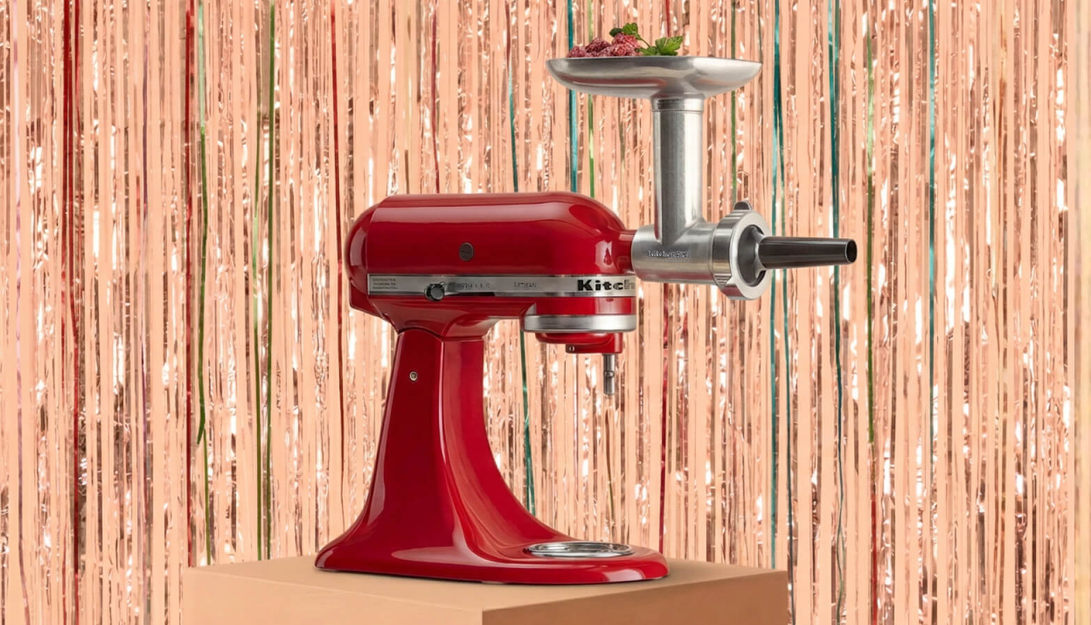 A KitchenAid® stand mixer with grinder attachment in front of a shiny decorative background