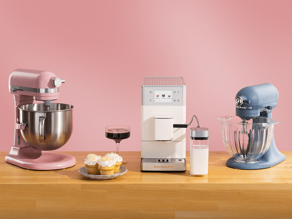 Pink KitchenAid® stand mixer, white espresso machine, and light blue stand mixer on a wood counter beside cupcakes and espresso
