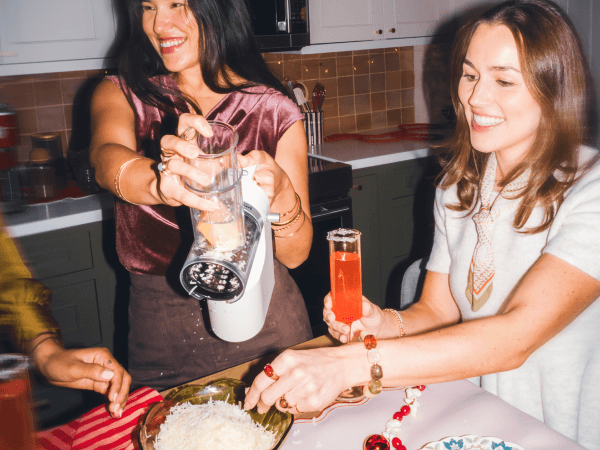 Woman grating cheese with a white KitchenAid® handheld grater beside a friend holding a cocktail at a holiday gathering