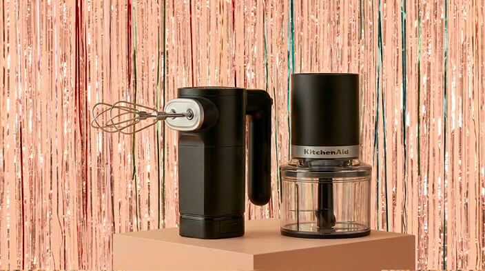 A KitchenAid® cordless hand mixer and food chopper in front of a shiny decorative background