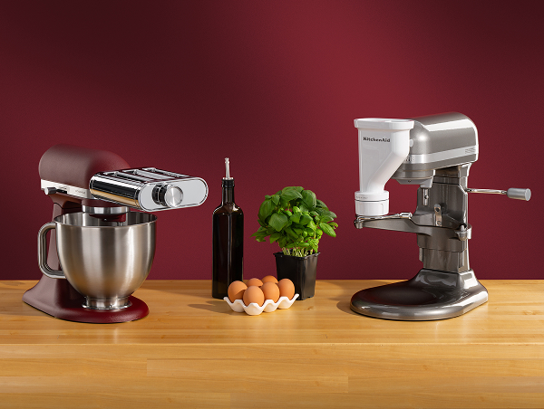 A red stand mixer with a pasta attachment and a silver stand mixer with a meat grinding attachment on a wooden counter with eggs, olive oil and basil