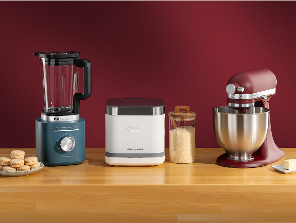 Blue KitchenAid® blender, white grain and rice cooker, and deep red stand mixer on a wooden counter with macarons and butter