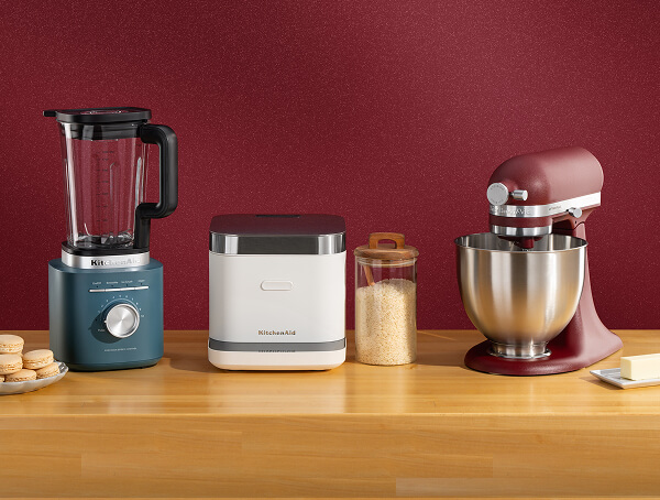 Blue KitchenAid® blender, white grain and rice cooker, and deep red stand mixer on a wooden counter with macarons and butter