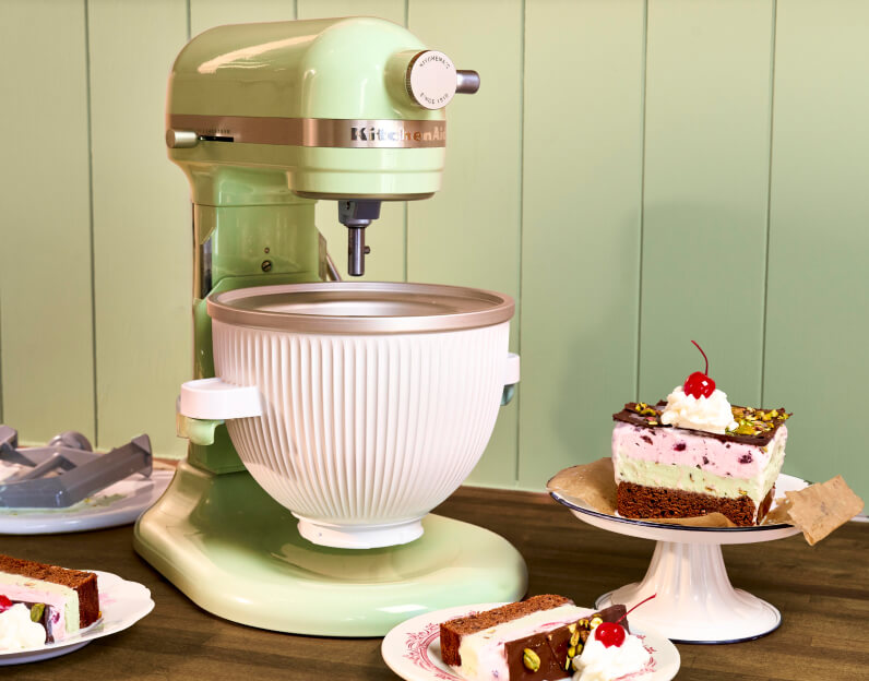 A KitchenAid® stand mixer with the Ice Cream maker attachment next to a slice of ice cream cake