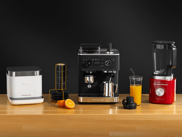 White KitchenAid® grain and rice cooker, black espresso machine, and red blender on a wood counter beside orange juice
