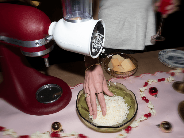 Red KitchenAid® stand mixer with a grater attachment shredding cheese into a bowl, beside a dish of cubed cheese on a holiday table