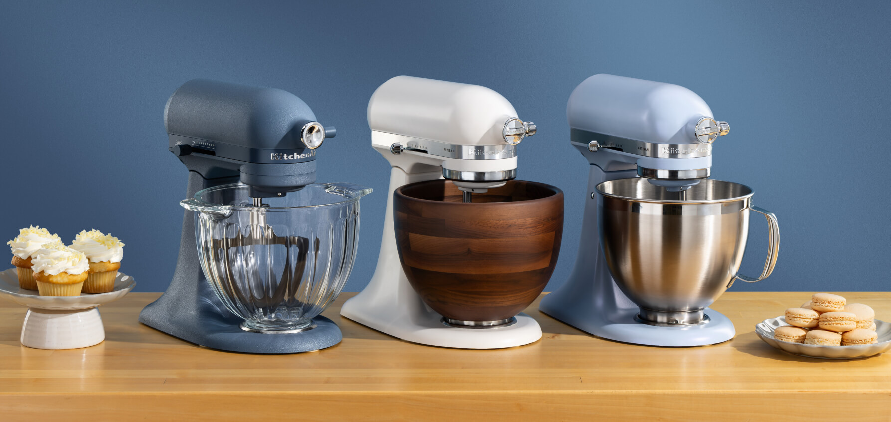 A Luminaire stand mixer, white stand mixer and blue stand mixer on a wooden counter with sweets on plates
