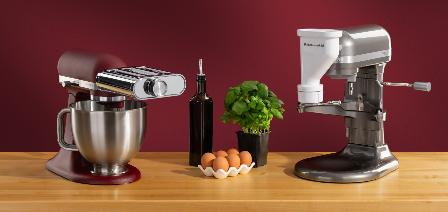 A red stand mixer with a pasta roller attachment and a gray stand mixer with a meat grinder attachment on a wooden counter with olive oil, eggs and a basil plant