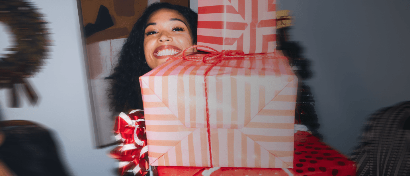 Woman smiling and holding wrapped holiday gifts in front of a Christmas tree