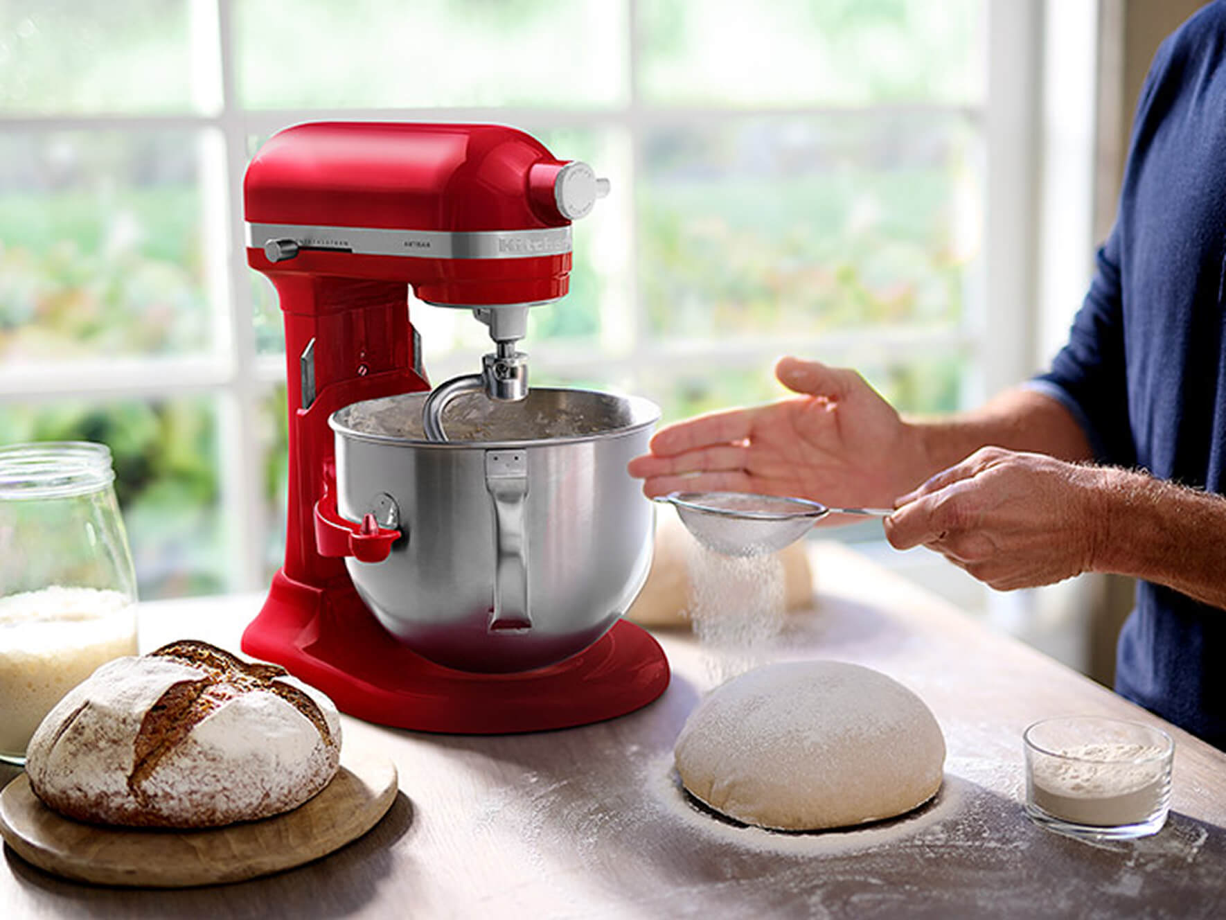 Person sifting flour over dough beside a red KitchenAid® bowl-lift stand mixer and a loaf of bread