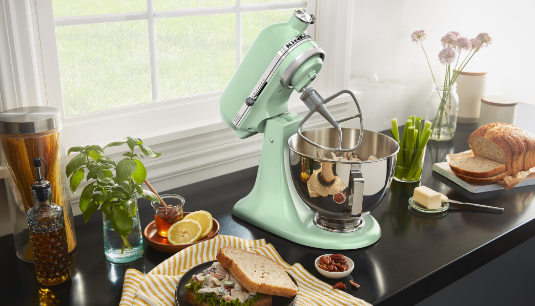 Green KitchenAid® stand mixer on a dark counter beside basil, lemons, jam, and sun-dried tomatoes