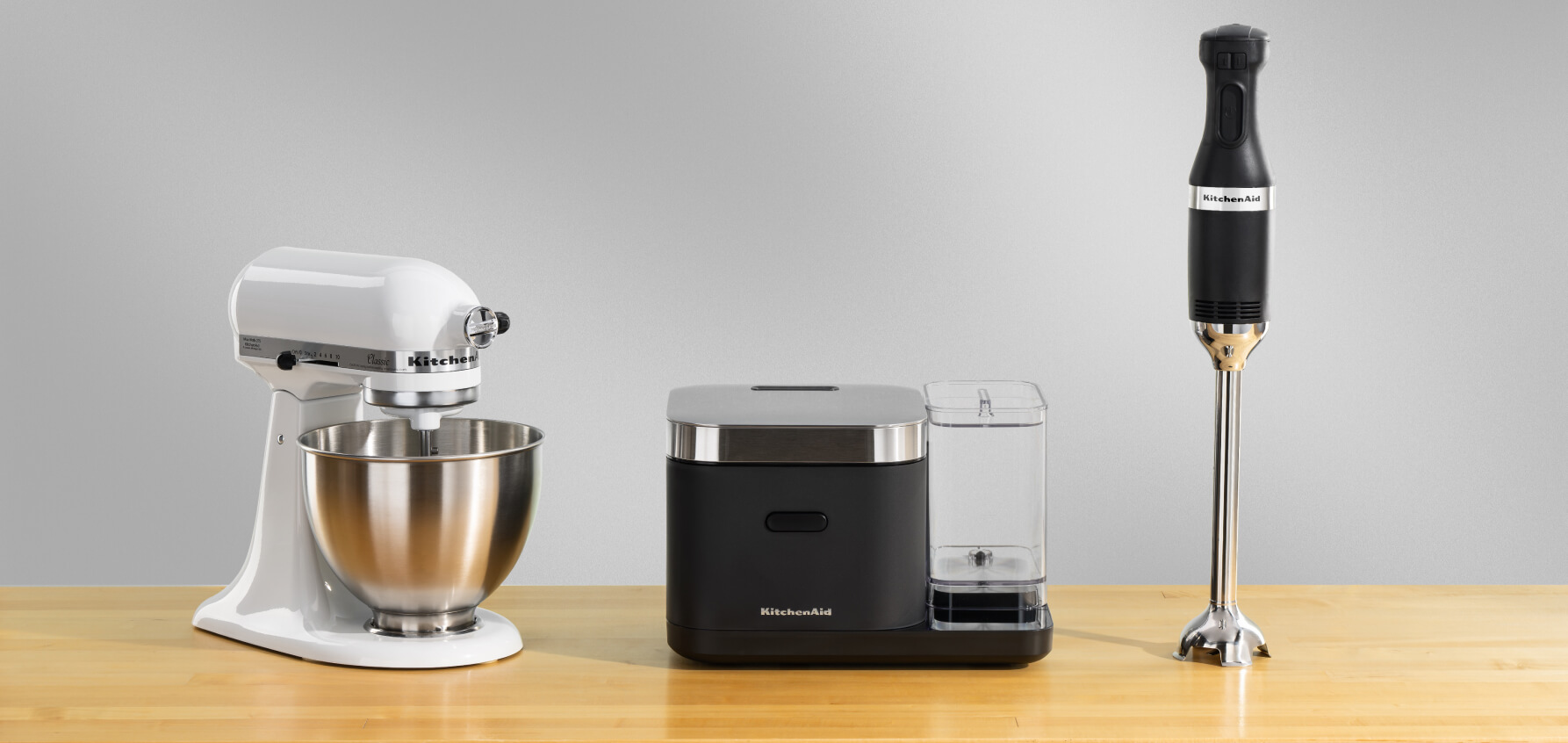 A white stand mixer, a black grain and rice cooker and a black immersion blender on a wooden counter