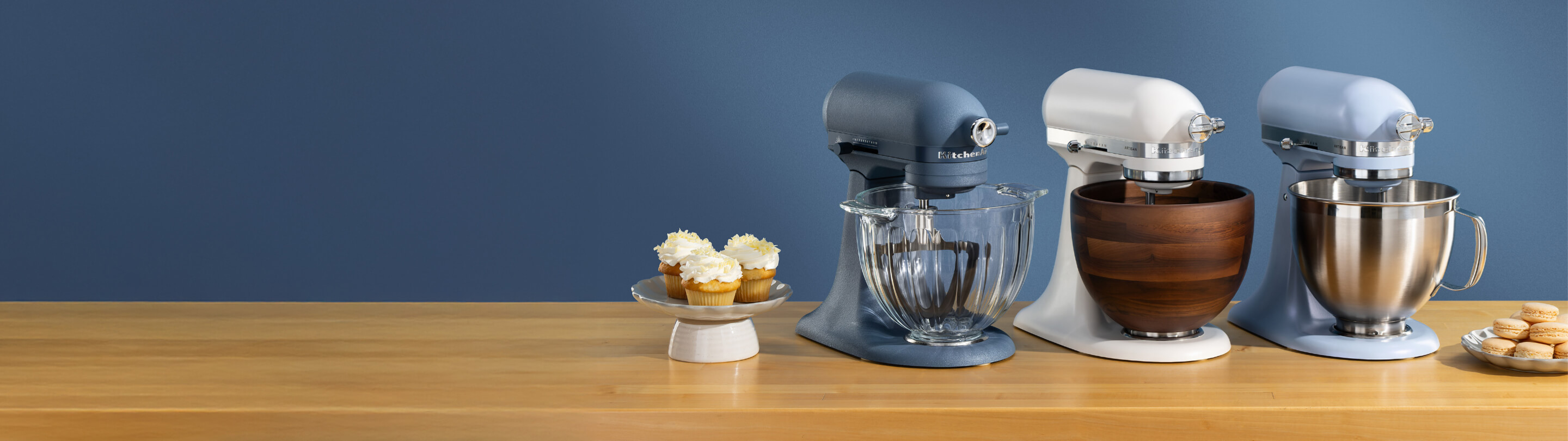 Blue, white and blue stand mixers on a wooden counter with different plates of pastries.