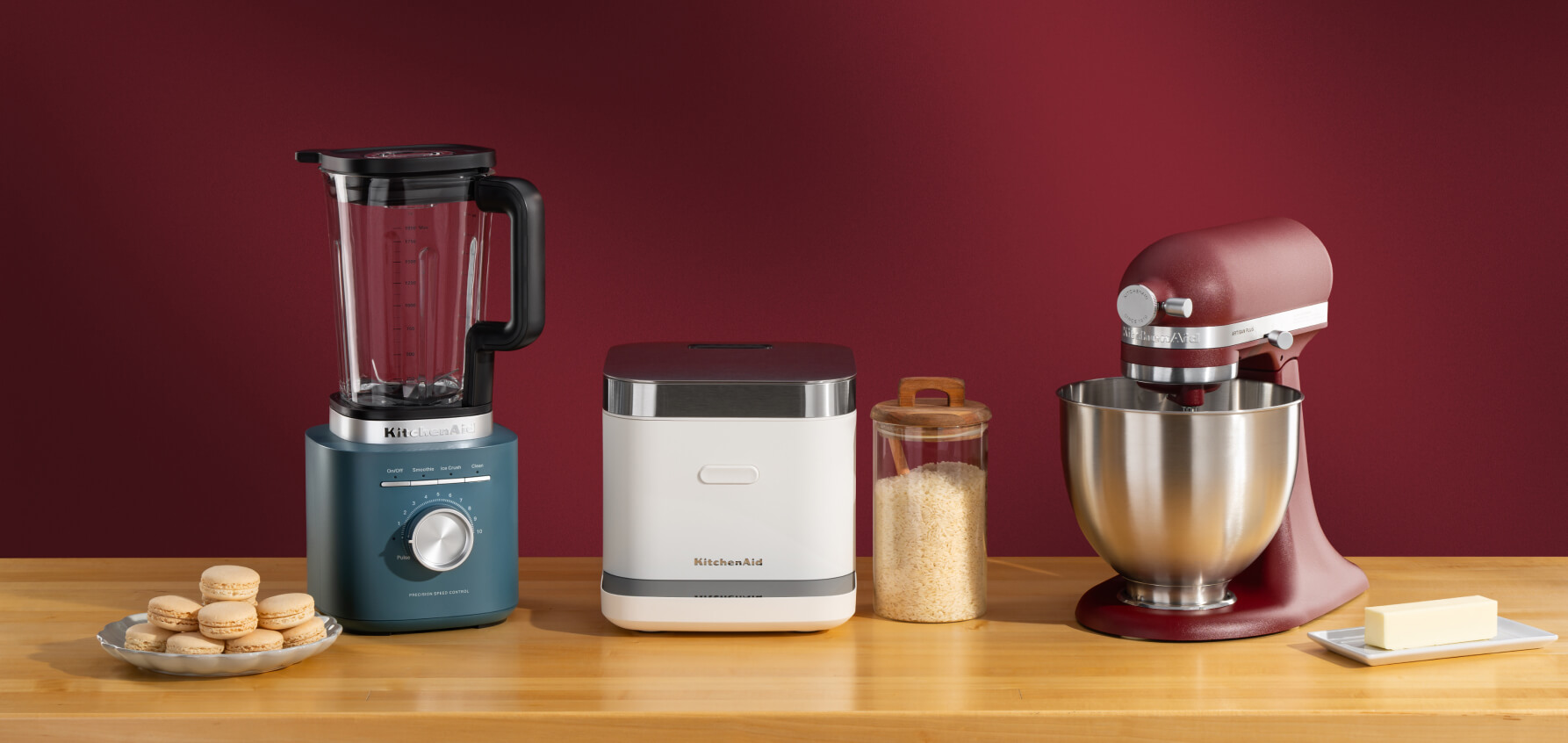 A blue blender, white grain and rice cooker and a red stand mixer on a counter with cookies and different ingredients