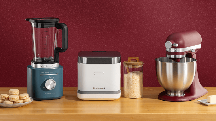 A blue blender, white grain and rice cooker and a red stand mixer on a counter with cookies and different ingredients