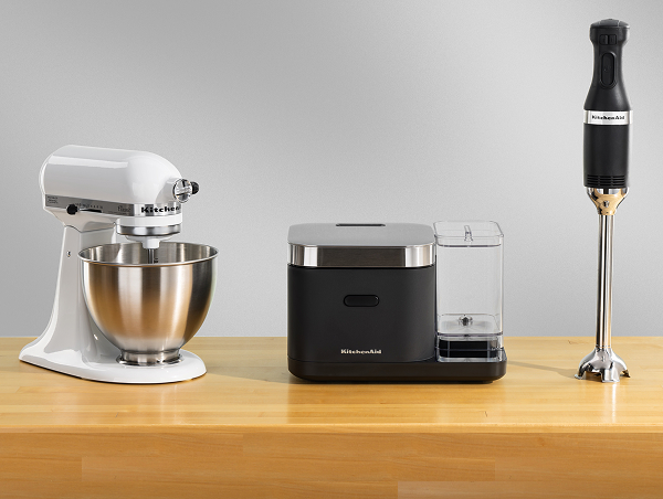 A white KitchenAid® stand mixer, a black grain and rice cooker and a black immersion blender on a wooden counter.