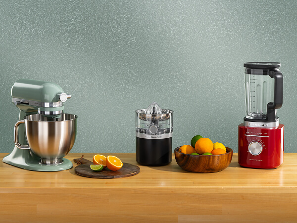 A teal stand mixer, black citrus juicer and red blender on a wooden counter with assorted citrus