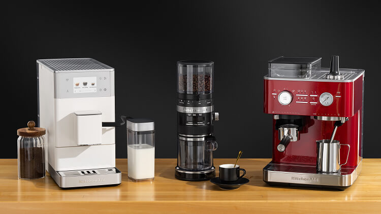 Various KitchenAid® coffee and espresso makers on a wooden counter surrounded by coffee ingredients