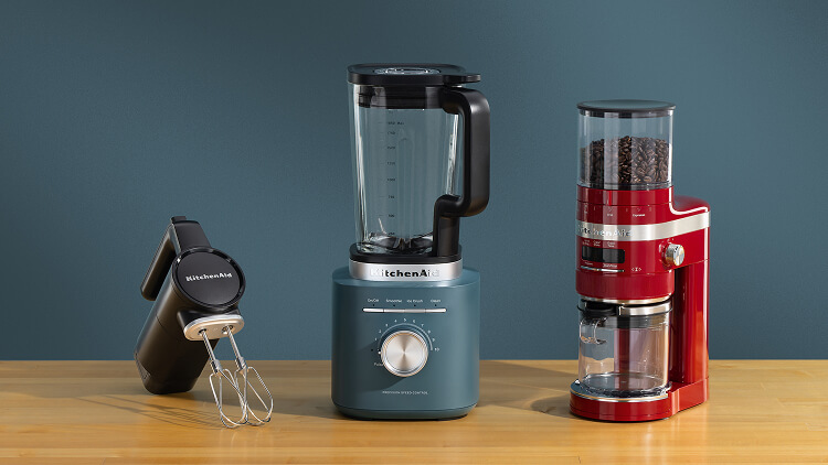 A black KitchenAid Go® Cordless hand mixer, a blue blender and a red bean grinder on a wooden counter.
