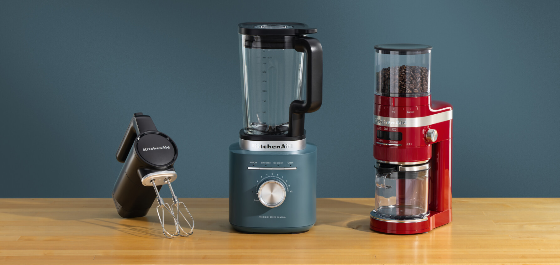 A black KitchenAid Go® cordless hand mixer, a blue blender and a red bean grinder on a wooden counter