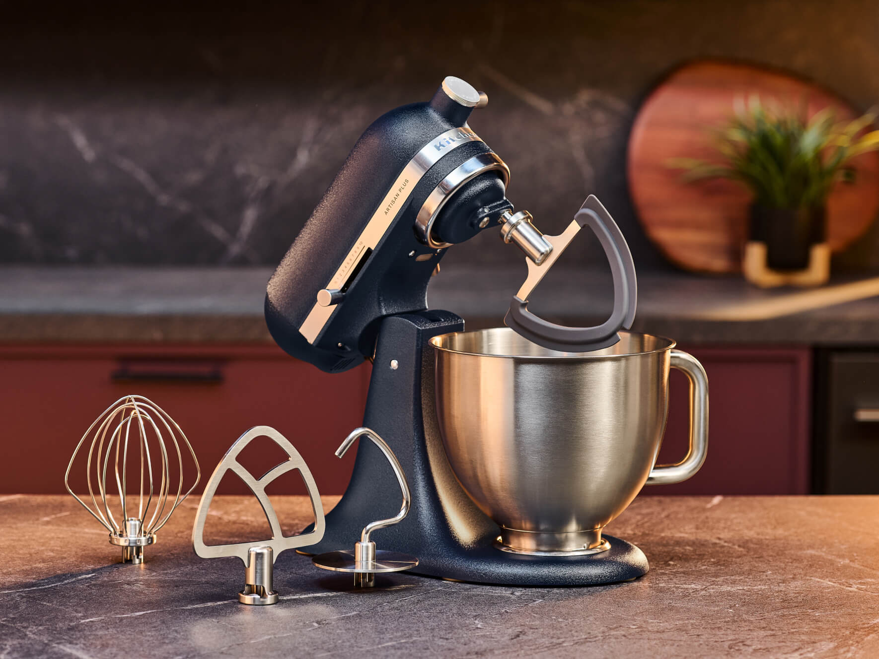 KitchenAid® Artisan Plus tilt-head stand mixer with a stainless steel bowl, beside whisk, beater, and dough hook attachments on a counter