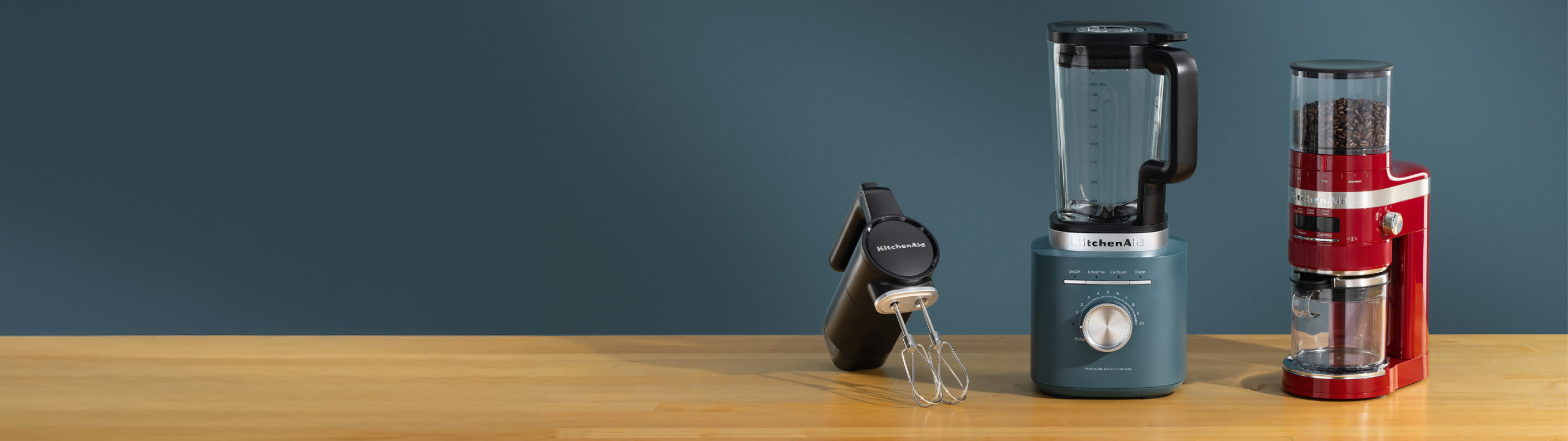 A black KitchenAid Go® cordless hand mixer, a blue blender and a red bean grinder on a wooden counter