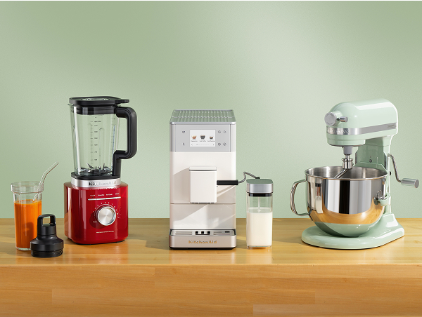 Red KitchenAid® blender, white espresso machine, and green stand mixer on a wooden counter with a glass of juice