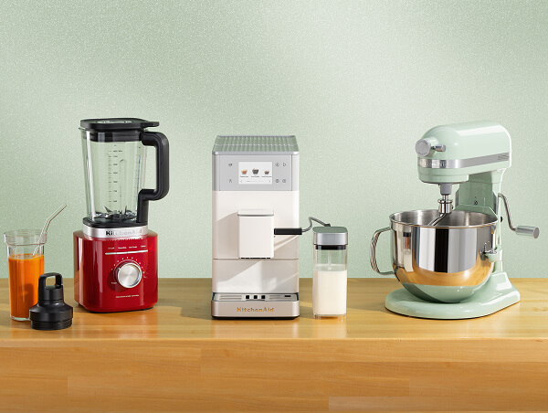 A red blender, white espresso machine and aqua stand mixer on a wooden counter with a smoothie in the blender jar.