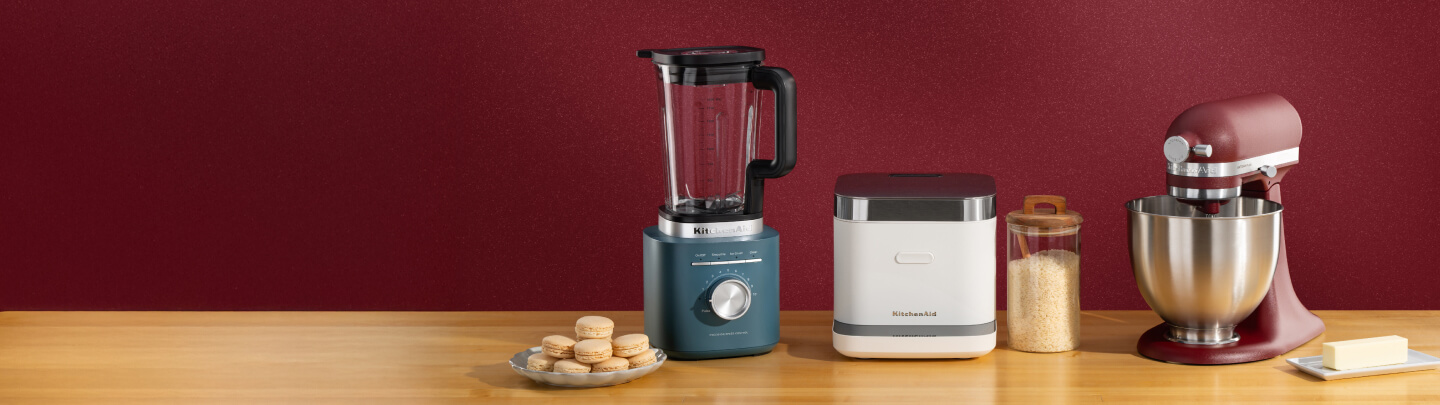 Blue KitchenAid® blender, white grain and rice cooker, and deep red stand mixer on a wooden counter with macarons and butter