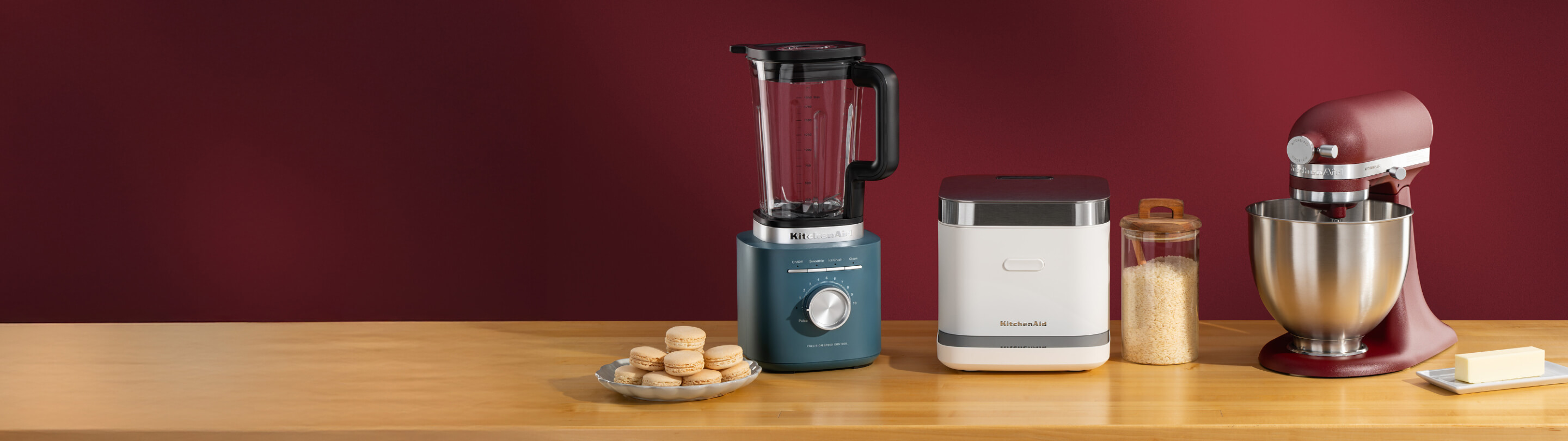 Blue KitchenAid® blender, white grain and rice cooker, and deep red stand mixer on a wooden counter with macarons and butter
