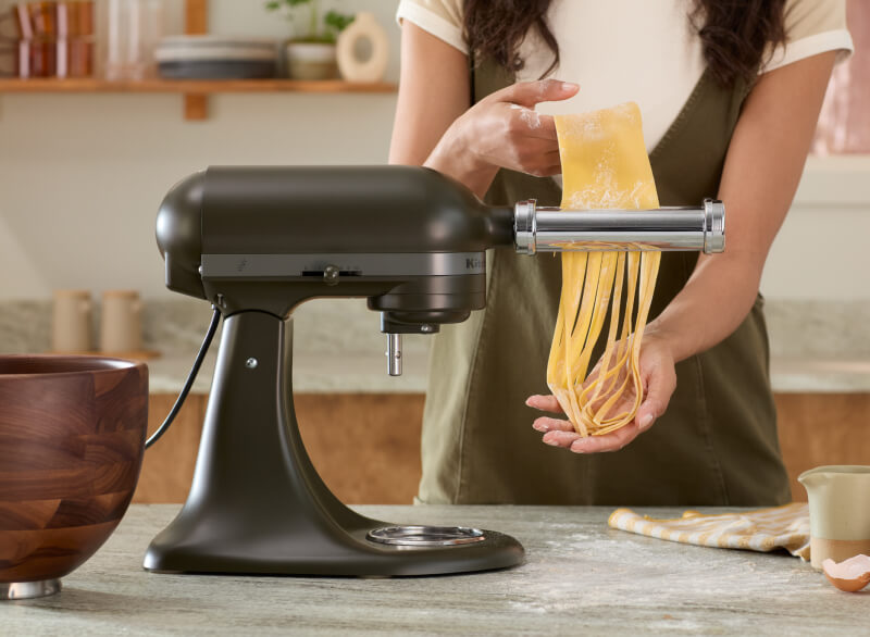 Person catching pasta noodles from a black KitchenAid® stand mixer with a pasta roller attachment, beside a walnut wood bowl