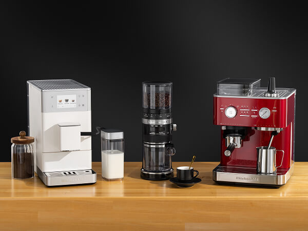 White KitchenAid® fully automatic espresso machine, black coffee grinder, and red semi-automatic espresso machine on a counter with milk and a mug