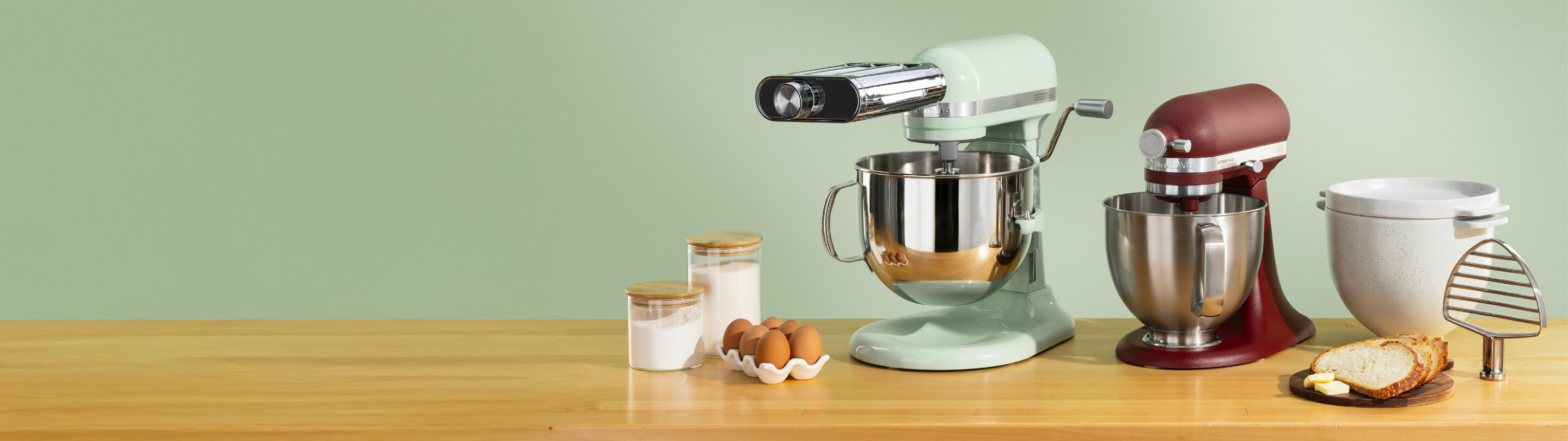 Green KitchenAid® stand mixer with a pasta roller attachment and burgundy stand mixer on a wood counter beside a ceramic bread bowl
