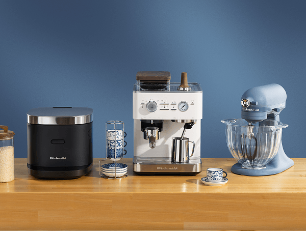 Black KitchenAid® rice cooker, white espresso machine, and blue Luminaire stand mixer with a glass bowl on a wood counter