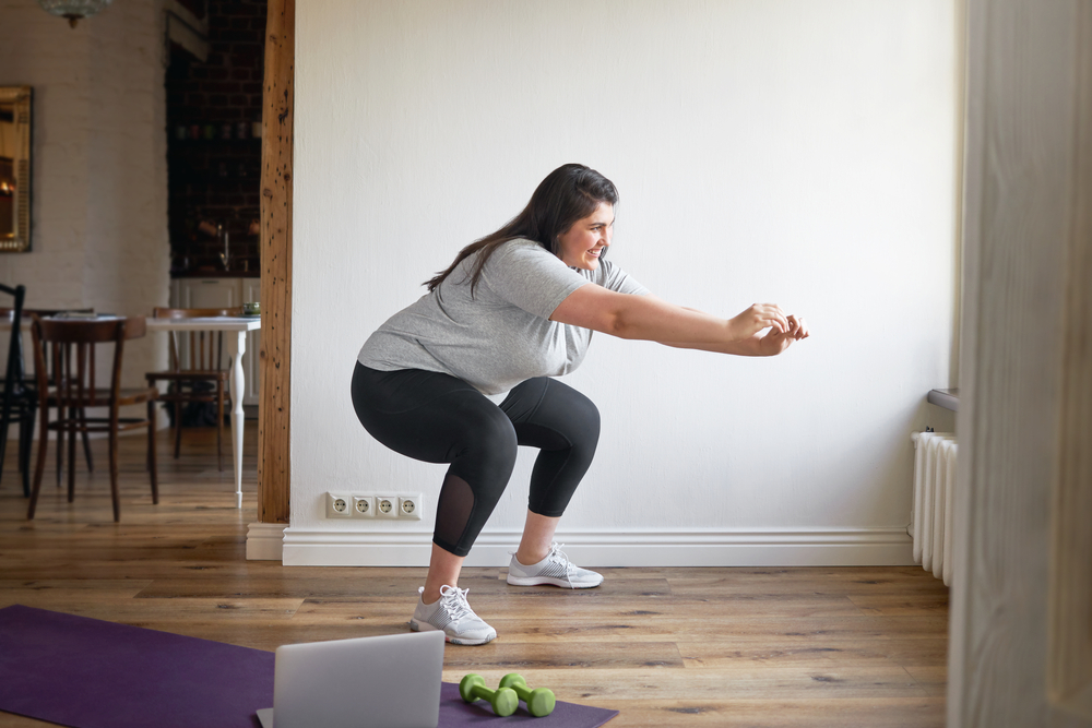 A woman in a grey t-shirt and black workout pants does a squat as she straightens her arms out in front of her.