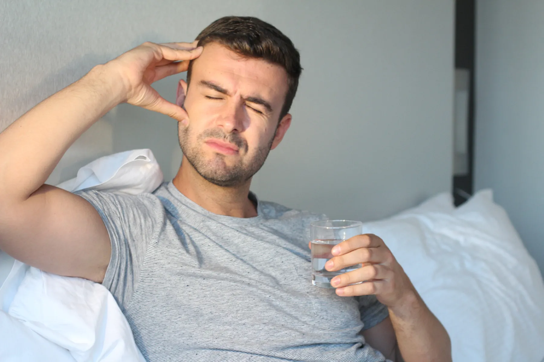 A man sits on his bed and holds his head in one hand and a glass of water in the other.