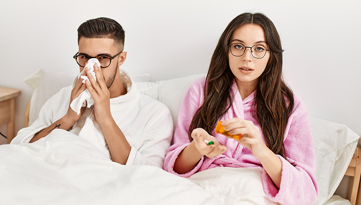 A couple sits in bed, the man is sneezing into a tissue and the woman pours medicine into her hand.