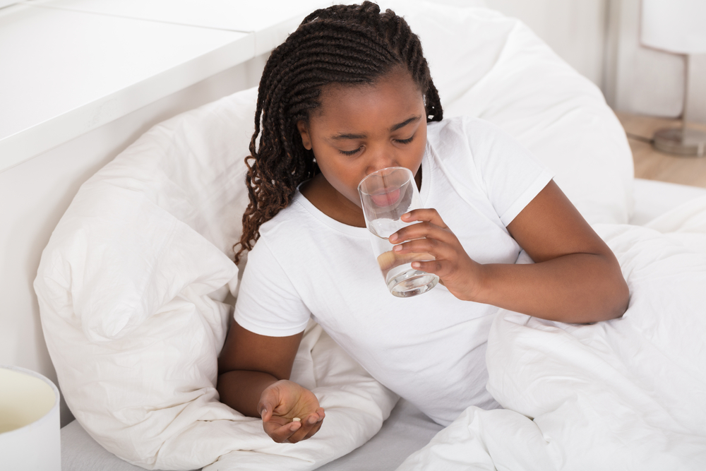 A woman in a bed with white linens drinks a glass of water while holding a pill in her hand.