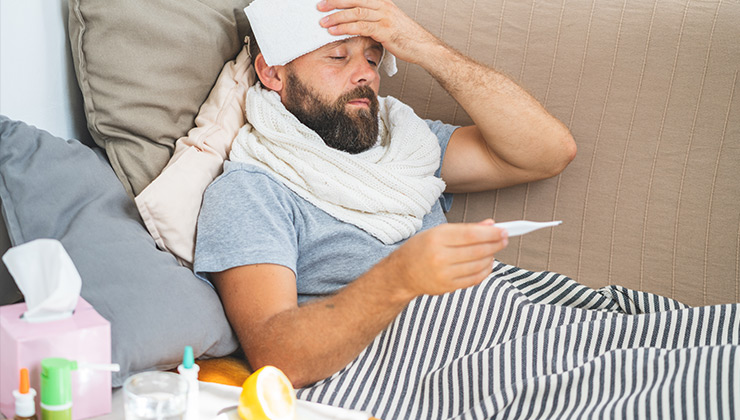 A sick man in bed holds a compress to his head and checks his temperature.