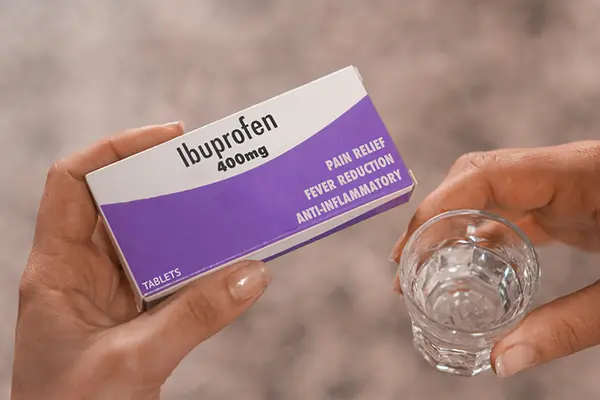 A person holds a box of ibuprofen and a glass of water.